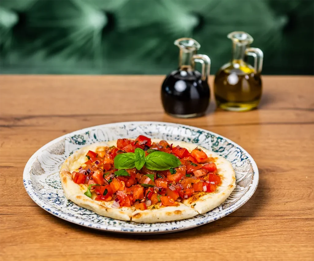 Frisch gebackenes Bruschetta-Pizzabrot belegt mit gewürfelten Tomaten, roten Zwiebeln und Basilikum auf einem Holztisch.