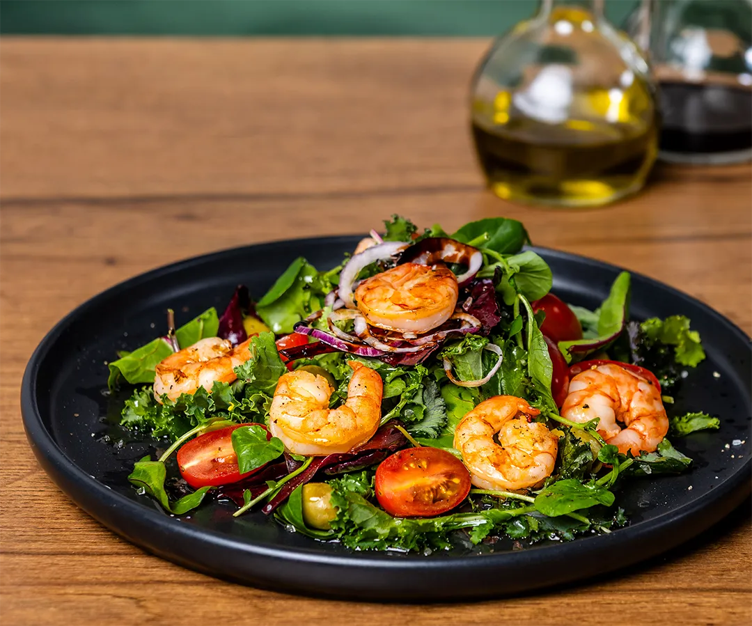 Frischer gemischter Blattsalat mit gebratenen Garnelen, halbierten Kirschtomaten, roten Zwiebeln und Oliven auf einem schwarzen Teller.