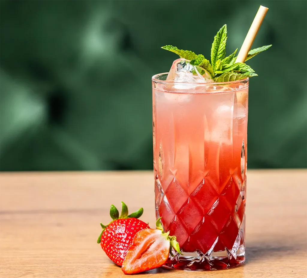 Kühle rote Erdbeer-Limonade im Kristallglas mit Eiswürfeln, Minze, Strohhalm und frischen Erdbeeren auf dem Holztisch.