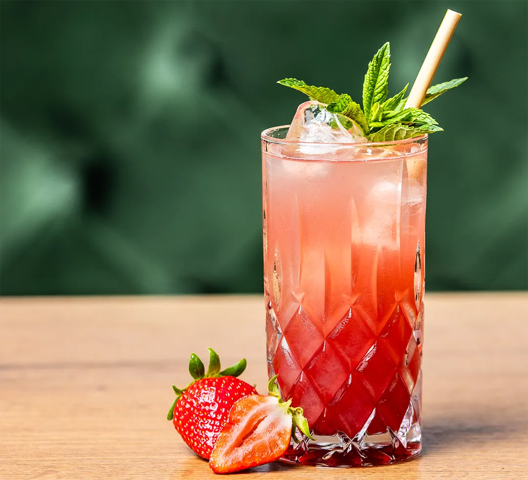 Kühle rote Erdbeer-Limonade im Kristallglas mit Eiswürfeln, Minze, Strohhalm und frischen Erdbeeren auf dem Holztisch.