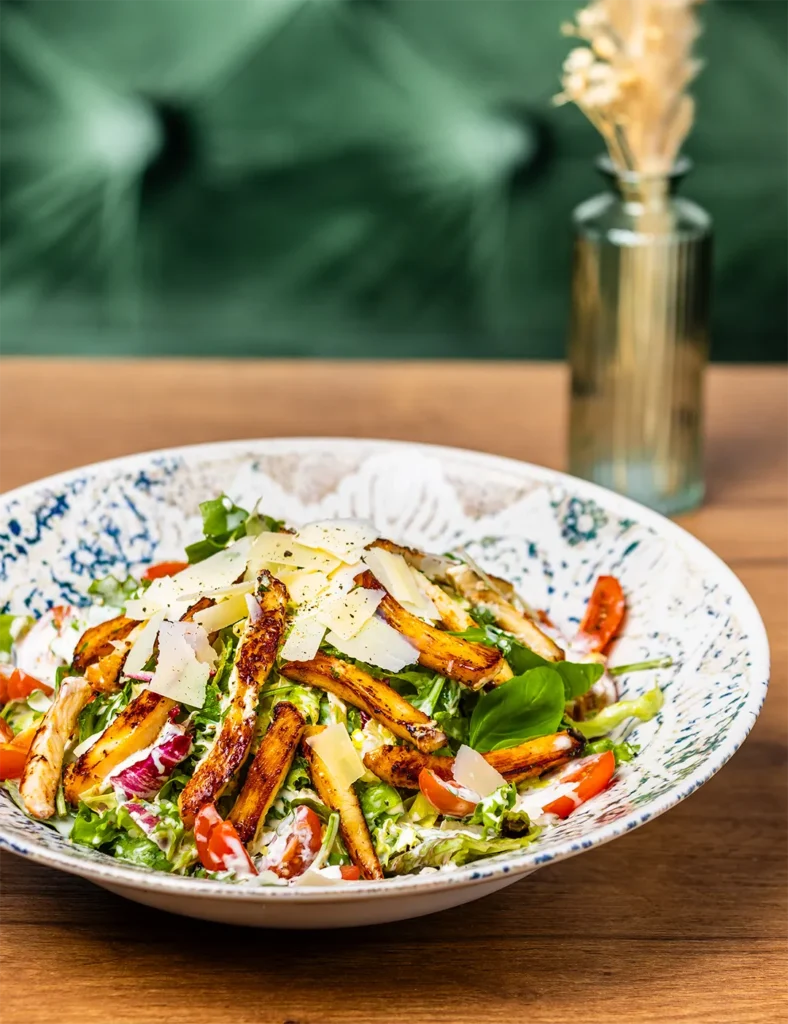 Caesar Salad mit gebratenen Hähnchenbruststreifen, Parmesan-Spänen, Kirschtomaten und hellem Dressing in einer tiefen Keramikschale.