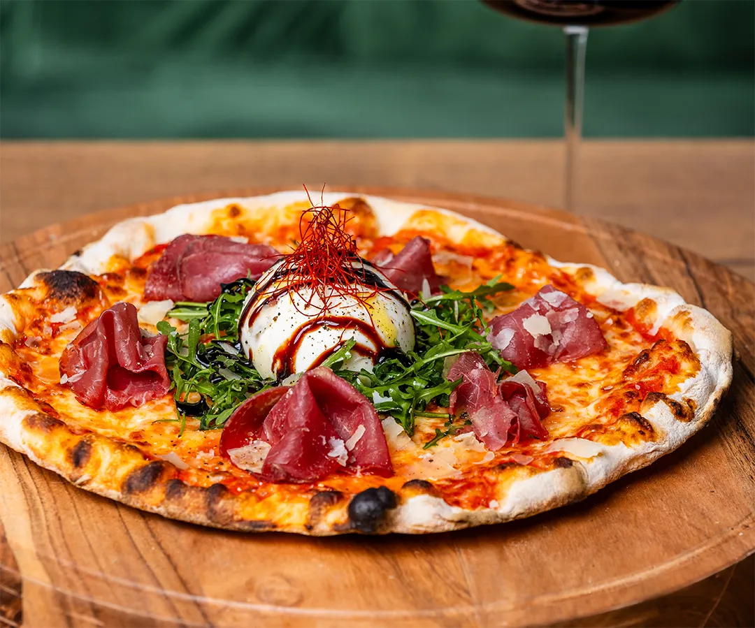 Italienische Steinofenpizza belegt mit Bresaola, Rucola, frischer Burrata, Balsamico-Creme und Chili-Fäden auf einem Holzbrett.