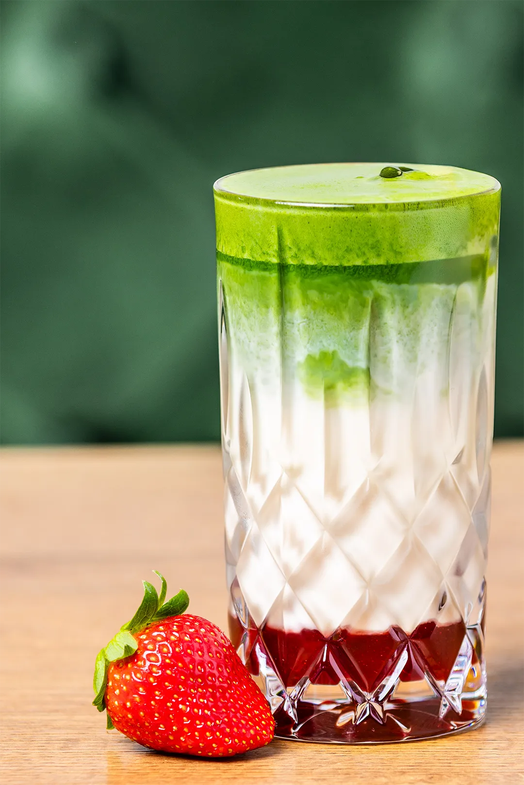 Eiskalter Strawberry Matcha Latte im Kristallglas mit roten Erdbeerpüree-Schichten, weißer Milch, grüner Matchaschicht und einer frischen Erdbeere.