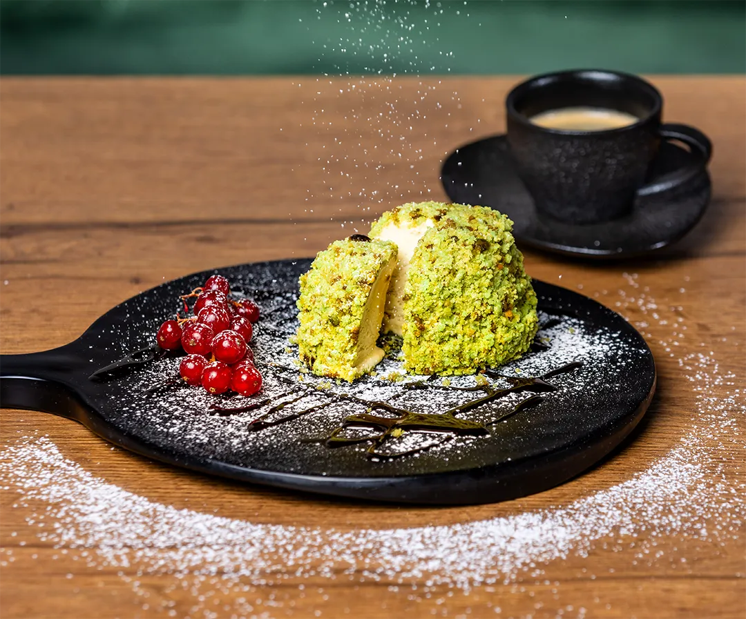 Pistazien-Tartufo-Eis auf einer schwarzen Servierplatte, bestreut mit Puderzucker, garniert mit roten Johannisbeeren und Schokosoße neben einer Tasse Espresso.