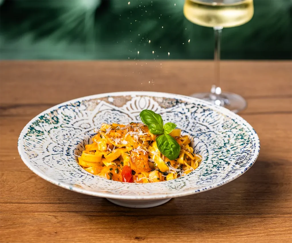 Frische Tagliatelle-Nudeln mit Garnelen, Parmesan und Basilikum auf einem verzierten Teller mit Weißweinglas.
