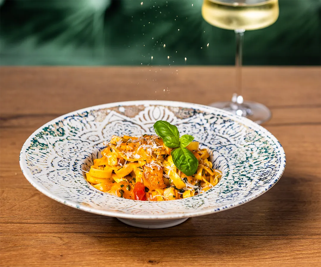 Frische Tagliatelle-Nudeln mit Garnelen, Parmesan und Basilikum auf einem verzierten Teller mit Weißweinglas.