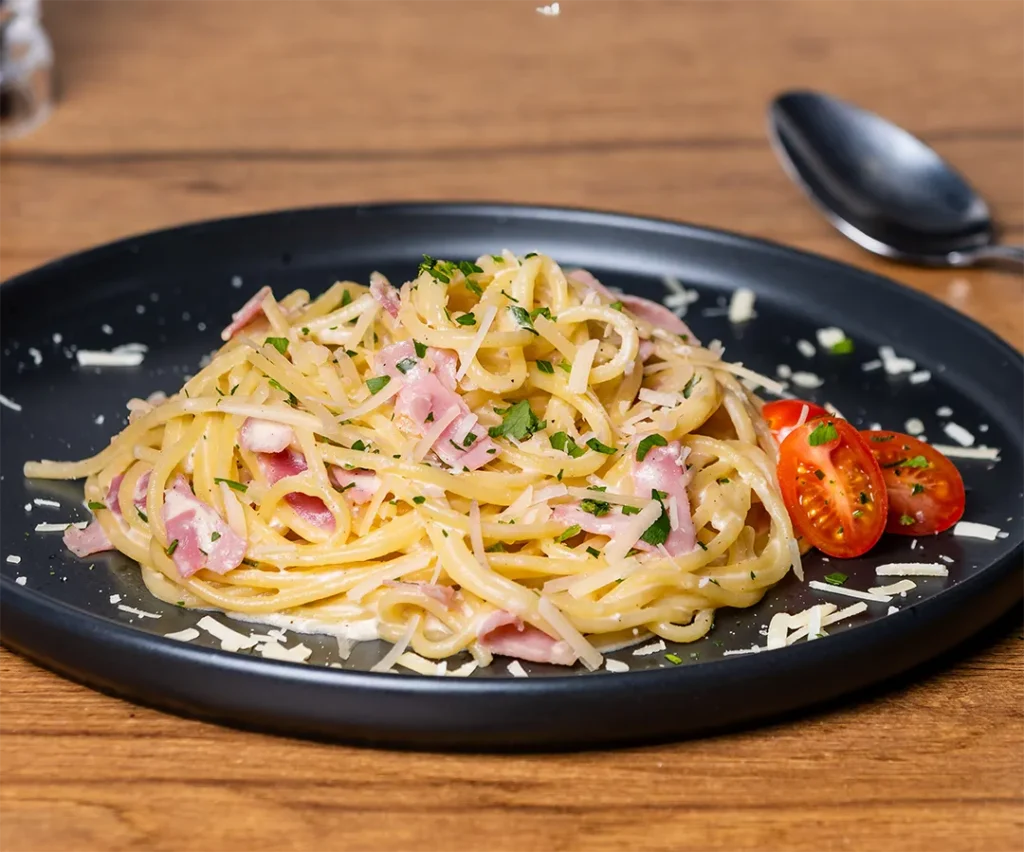 Spaghetti Carbonara mit Kochschinken, gehobeltem Parmesan, Petersilie und Kirschtomaten auf einem schwarzen Teller.