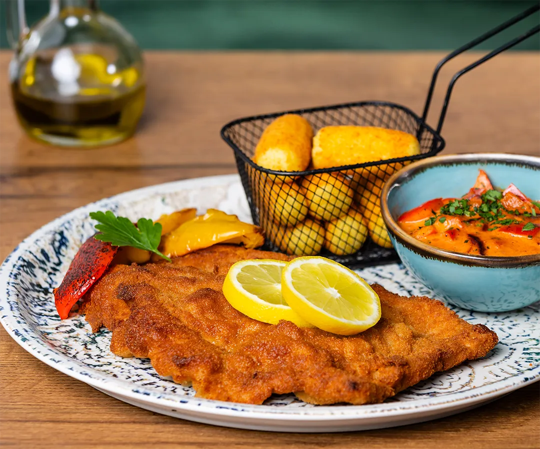 Goldbraun paniertes Schnitzel mit Zitronenscheiben, Kroketten im Servierkörbchen und Würzig-cremiger Soße auf einem Teller.